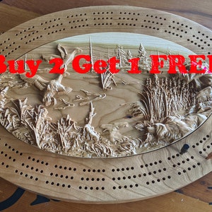 May include: Wooden cribbage board with a detailed, carved nature scene featuring ducks, reeds, and trees. The board has a series of holes for pegging and includes the text "Buy 2 Get 1 FREE!!!". A deck of cards is visible.