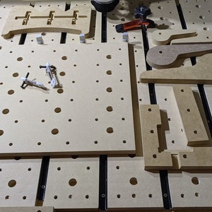 May include: A close-up of a wooden work surface with a grid of holes. There are several pieces of wood cut into different shapes, including a handle and a bracket. The surface is covered in a layer of sawdust.