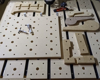Puede incluir: Primer plano de una superficie de trabajo de madera con una cuadrícula de agujeros. Hay varias piezas de madera cortadas en diferentes formas, incluyendo un mango y un soporte. La superficie está cubierta por una capa de aserrín.