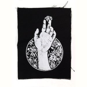 Może przedstawiać: Czarno-biała naszywka haftowana przedstawiająca rękę sięgającą w górę z małym owadzie na czubku palca. Ręka jest otoczona stylizowanymi kwiatami i liśćmi.