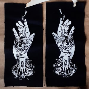 May include: Two black fabric patches with a white hand design, each featuring an eye in the palm. The hands are reaching upwards, with flowing details at the wrist. A skull and bone are above the patches.