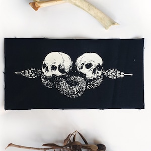 Puede incluir: Un parche negro con un gráfico blanco de dos calaveras y una serpiente. Encima del parche hay dos huesos y debajo una ramita de hojas secas. El parche tiene un borde sin rematar.