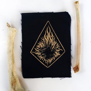 May include: A black fabric patch with a gold flame design inside a diamond shape. Two bone fragments are positioned on either side of the patch against a white background. The patch has a raw edge.