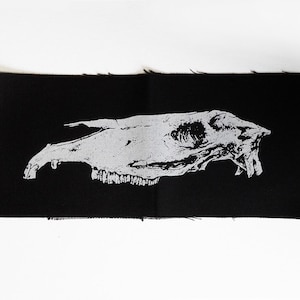 Könnte beinhalten: Ein schwarzer Stoff-Patch mit einer weißen Umrandung eines Tierschädels. Der Schädel ist nach rechts gerichtet und hat sichtbare Zähne.
