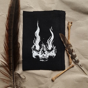 May include: Black fabric patch featuring a white skull design. The skull's eyes are replaced with flames.
