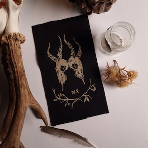 May include: A black fabric patch with a gold illustration of two horned skulls. The design includes a floral wreath and two runes. The patch is rectangular and made of woven material. The overall aesthetic is gothic and mystical.