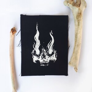 K&ouml;nnte beinhalten: Schwarzer Stoff-Patch mit einem wei&szlig;en Totenkopf-Design. Der Sch&auml;del hat Flammen als Augen.