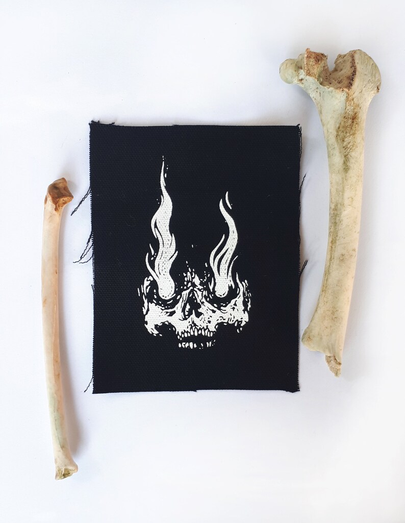 K&ouml;nnte beinhalten: Schwarzer Stoff-Patch mit einem wei&szlig;en Totenkopf-Design. Der Sch&auml;del hat Flammen als Augen.