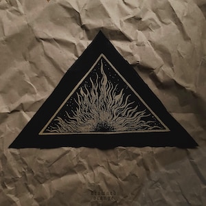 May include: Black triangular patch with a gold sunburst design. The sunburst has long, wavy lines emanating from a central point. The patch is on a brown, crumpled paper surface. The text "drowned orange" is at the bottom.