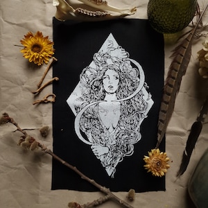 May include: Black patch featuring a white illustration of a woman with flowing hair, set within a diamond shape. A crescent moon design frames the woman. The patch is surrounded by dried flowers, feathers, and bones.