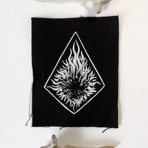 Pode incluir: Um patch preto com um design branco em forma de diamante com um sol ou chama estilizada. Dois fragmentos ósseos são posicionados acima e abaixo do patch em um fundo branco. O patch tem bordas cruas.