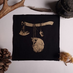 May include: Black square patch with a gold illustration of a skull, axe, and other objects hanging from a wooden beam. The patch has a raw edge and is likely made of fabric.
