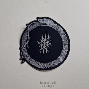 May include: A black and white sticker with a snake curled into a circle. The snake's head is facing the left side of the circle. Inside the circle is a black background with a white symbol that looks like a series of vertical and horizontal lines.