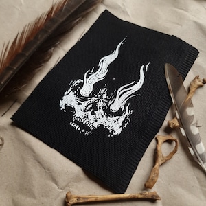 K&ouml;nnte beinhalten: Ein schwarzer Stoff-Patch mit einem wei&szlig;en Totenkopf-Design. Der Sch&auml;del hat Flammen, die aus seinen Augenh&ouml;hlen kommen.