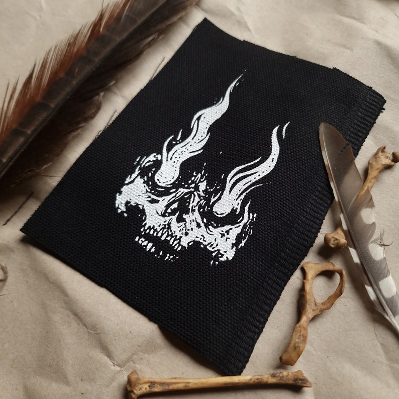 K&ouml;nnte beinhalten: Ein schwarzer Stoff-Patch mit einem wei&szlig;en Totenkopf-Design. Der Sch&auml;del hat Flammen, die aus seinen Augenh&ouml;hlen kommen.