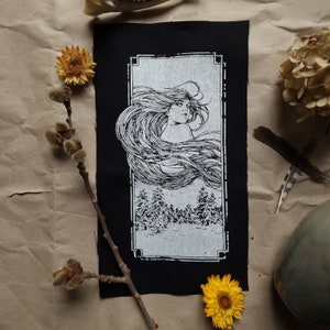 May include: Black rectangular patch with a white illustration of a woman with flowing hair above a forest scene. The patch is framed with a decorative border. Dried flowers and a branch with buds are arranged around the patch.