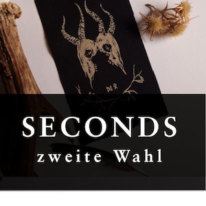 Peut inclure: Écusson rectangulaire noir avec une illustration dorée de deux crânes de chèvre. Le patch porte les mots "SECONDS" et "zweite Wahl" imprimés en blanc. L'image comprend une fleur séchée et un morceau de bois de cerf.
