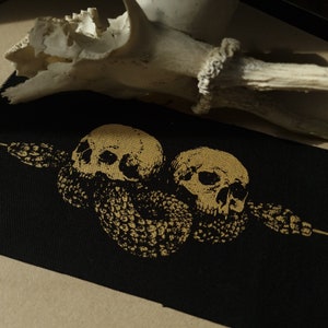 May include: Black fabric with a gold skull design. Two skulls are centered, with a snake-like design wrapping around them. The skulls and snake are in a gold color. A white antler is in the background.