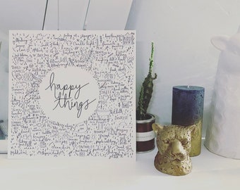 Puede incluir: Una impresión en blanco y negro con las palabras "happy little things" rodeadas de una variedad de ilustraciones dibujadas a mano. La impresión se muestra en un estante blanco con un cactus, una vela y una figurita de animal dorada.