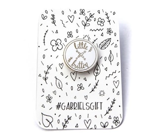 May include: White enamel pin with a gold outline and the words "Little Button" and a small cross inside. The pin is on a white card with a black and white doodle pattern and the text "#GABRIELSGIFT".