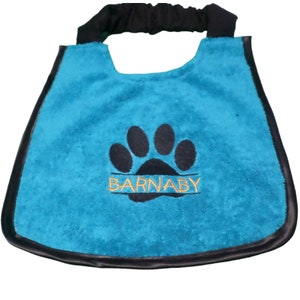 Puede incluir: Un bandana para perro azul turquesa con una huella de pata negra y el nombre "BARNABY" bordado en hilo naranja. El bandana tiene una correa ajustable negra.