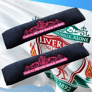 Könnte beinhalten: Zwei schwarze Kopfstützen für Autositze mit rosa Stickerei. Die Stickerei zeigt ein Wappen mit den Worten "You'll Never Walk Alone".