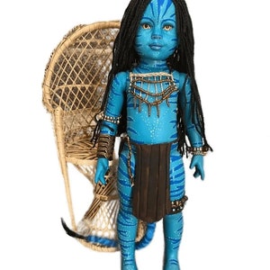K&ouml;nnte beinhalten: Eine blaue Puppe mit einer langen schwarzen Per&uuml;cke, die einen braunen Rock und Perlen-Schmuck tr&auml;gt. Die Puppe steht vor einem Korbstuhl.