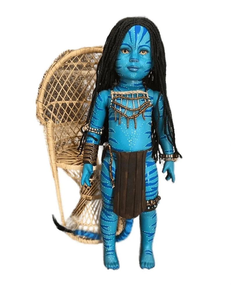 K&ouml;nnte beinhalten: Eine blaue Puppe mit einer langen schwarzen Per&uuml;cke, die einen braunen Rock und Perlen-Schmuck tr&auml;gt. Die Puppe steht vor einem Korbstuhl.