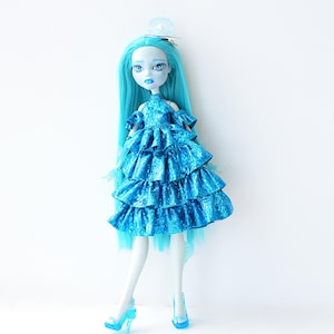 May include: A doll with long, light blue hair and matching blue eyes. The doll wears a tiered, ruffled blue dress and blue high heels. A small, decorative hair clip adorns the doll's hair. The doll's skin is pale blue, and the overall aesthetic is fantasy-themed.