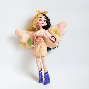K&ouml;nnte beinhalten: Eine Puppe mit blonden und schwarzen Haaren, die ein beiges Kleid mit einem rosa Oberteil und Schmetterlingsfl&uuml;geln tr&auml;gt. Die Puppe hat blaue Augen und tr&auml;gt lila Schuhe.