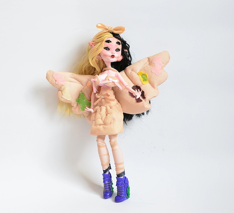 K&ouml;nnte beinhalten: Eine Puppe mit blonden und schwarzen Haaren, die ein beiges Kleid mit einem rosa Oberteil und Schmetterlingsfl&uuml;geln tr&auml;gt. Die Puppe hat blaue Augen und tr&auml;gt lila Schuhe.