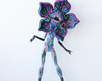 Könnte beinhalten: Eine humanoide Figur mit einem großen, blumenförmigen Kopf mit violetten, blauen und pinken Blütenblättern und einem zentralen Mund mit spitzen weißen Zähnen. Der Körper ist hellgrau mit blauen und pinken Ader-Mustern, schwarzen Gliedmaßen und Krallen.