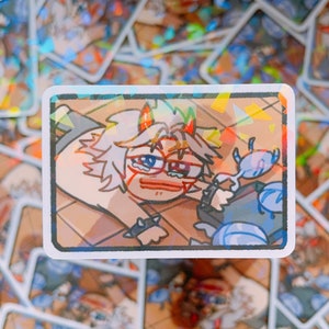 Peut inclure: Un autocollant rectangulaire représentant un personnage de dessin animé avec des cornes rouges, des lunettes et des larmes. Le personnage tend la main vers un objet bleu. L'autocollant a une finition holographique et est entouré d'autocollants similaires.