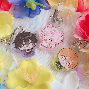 May include: Three anime-style acrylic charms with silver keychains, each depicting a different character. The charms are surrounded by artificial flowers in various colours. The characters have black, pink, and orange hair.