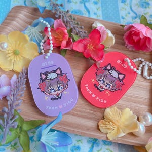 May include: Two keychains, one purple and one red, each featuring a cartoon character with cat ears and bows. The text "07.09" is at the top, and "YOON JIGU" is at the bottom. Both keychains have a white beaded chain.