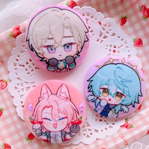 Puede incluir: Tres insignias redondas de felpa con diseños de personajes de estilo anime. Cada insignia tiene un fondo rosa y una ilustración de personaje diferente con rasgos faciales detallados y acentos coloridos. Las insignias están sobre un mantel blanco.