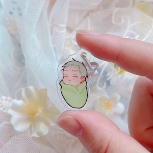 May include: A clear acrylic keychain featuring a cartoon illustration of a baby wrapped in a green blanket. The baby has closed eyes and light brown hair. The keychain has a silver-colored ring.