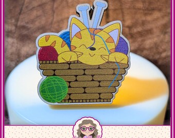 May include: A wooden pin featuring a yellow tabby cat with orange stripes sitting inside a brown woven basket. The cat is surrounded by colorful balls of yarn in red, blue, green, and purple, with two light blue knitting needles behind its head.