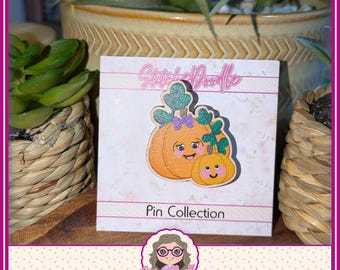 May include: A wooden pin featuring two orange pumpkins with smiling faces, green glittery leaves, and a small purple bow. The pin is attached to a white display card with the text StitchDoodle and Pin Collection printed in black and pink.
