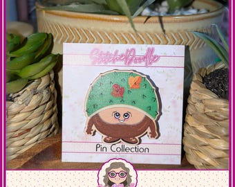 May include: A wooden pin featuring a cartoon character with a green, spiky, dome-shaped hat adorned with small orange and red leaves. The character has a tan face, large eyes, a smiling mouth, and a brown, rounded body on a display card.