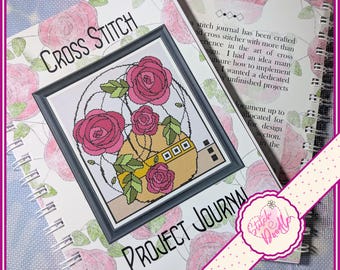 May include: A spiral-bound cross stitch project journal with a white cover featuring a pink rose pattern. The cover displays the text Cross Stitch Project Journal and a central illustration of pink roses in a yellow vase inside a dark frame.