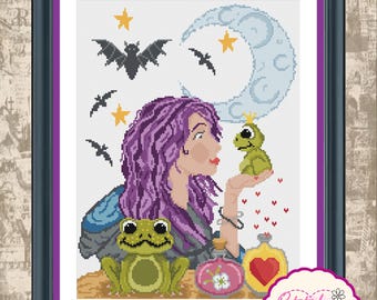 May include: A framed cross-stitch pattern featuring a person with purple hair holding a small green frog wearing a crown. Below, a larger green frog sits near two potion bottles. The background includes a light blue crescent moon, yellow stars, and black bats.
