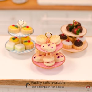 Puede incluir: Tres soportes de dos niveles en miniatura con pasteles de tama&ntilde;o casa de mu&ntilde;ecas. Los soportes tienen asas de metal dorado y platos con rayas azules y blancas, rosa s&oacute;lido y crema s&oacute;lido. El texto dice: Pastry sets available.