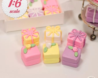 May include: A set of miniature gift boxes in various pastel colors, including pink, yellow, purple, and white. Each box is decorated with a bow and a small flower. The boxes are arranged on a white surface. The text "1:6 scale" is visible in the upper left corner of the image.