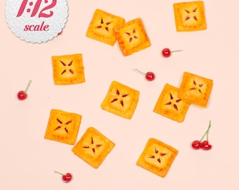 May include: Miniature square pies with a golden crust and a star-shaped filling, scattered on a light pink surface. Small red cherries are also scattered around the pies. The image includes the text "1:12 scale" in red.
