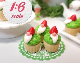 May include: Miniature cupcakes with green frosting, red hats, and white beards, resembling garden gnomes. The cupcakes sit on a green doily. The image includes the text "1:6 scale" in a white circle.