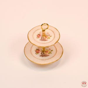 Puede incluir: Un soporte de servicio en miniatura de dos niveles con platos de color crema, un delicado patr&oacute;n de puntos rosas, bordes festoneados dorados y un dise&ntilde;o de ramo floral central. El soporte est&aacute; unido por una varilla central de metal dorado.