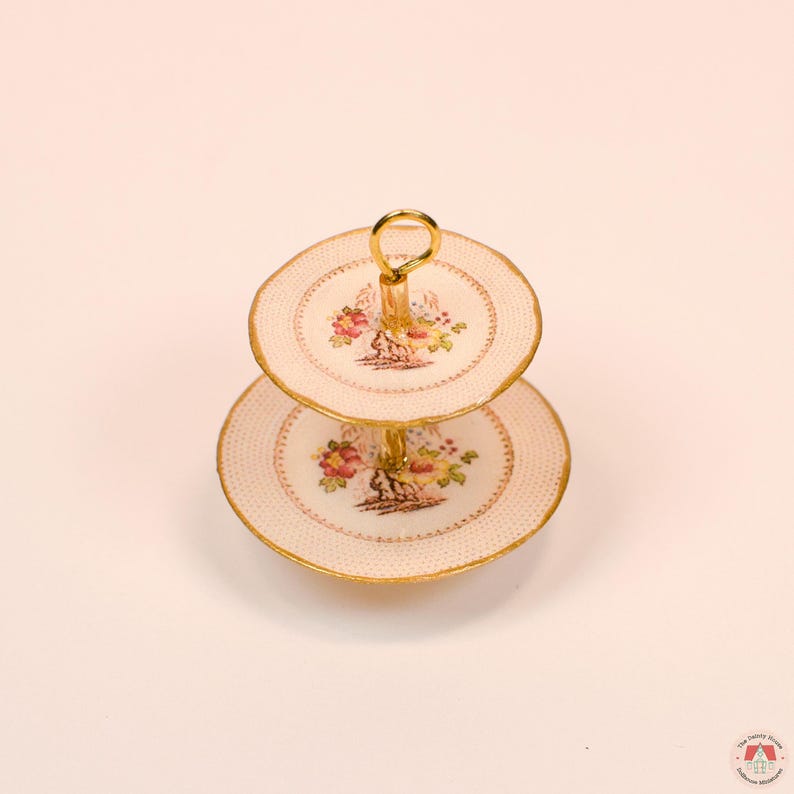 Puede incluir: Un soporte de servicio en miniatura de dos niveles con platos de color crema, un delicado patr&oacute;n de puntos rosas, bordes festoneados dorados y un dise&ntilde;o de ramo floral central. El soporte est&aacute; unido por una varilla central de metal dorado.