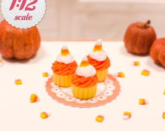 May include: Three miniature cupcakes in yellow liners with orange frosting and a candy corn topper, arranged on a scalloped paper doily. The scene includes scattered candy corn pieces and two small pumpkins, with a 1:12 scale label visible.