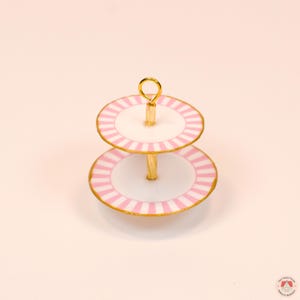 Puede incluir: Un soporte de dos niveles con platos circulares blancos, bordes de rayas rosas y acabados dorados. Los niveles est&aacute;n conectados por un poste central dorado que termina en una peque&ntilde;a anilla en la parte superior para colgarlo.
